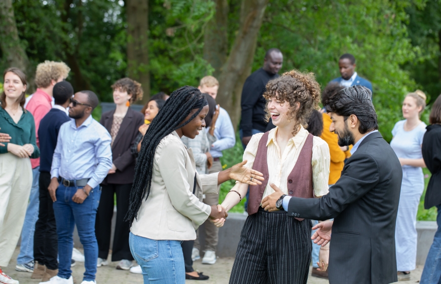 People from different nationalities standing together outside underneath trees and shaking hands, laughing.